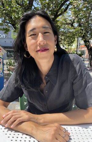 A long-haired Asian woman sits at a table in the sun in front of some trees.