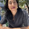 A long-haired Asian woman sits at a table in the sun in front of some trees.